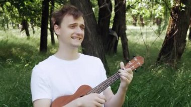 Genç adam bir tarlada ukulele çalıyor ve enerjik bir şekilde şarkı söylüyor. Genç hippi doğada ukulele ya da gitar çalıp şarkı söylüyor.. 