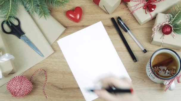 女性の手書きは 贈り物 包装紙 赤いクリスマスの装飾と木製のテーブルにギフトリストを書きます Xmasとハッピーニューイヤーの構成 フラットレイ トップビュー — ストック動画