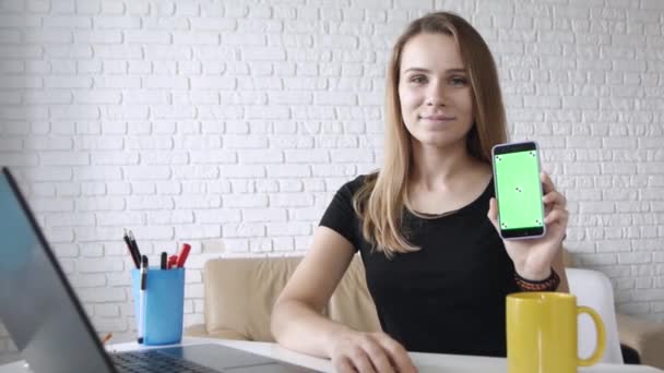 年轻的白种人长发女孩的肖像微笑着显示绿色屏幕的智能手机相机左手在家庭办公室 — 图库视频影像