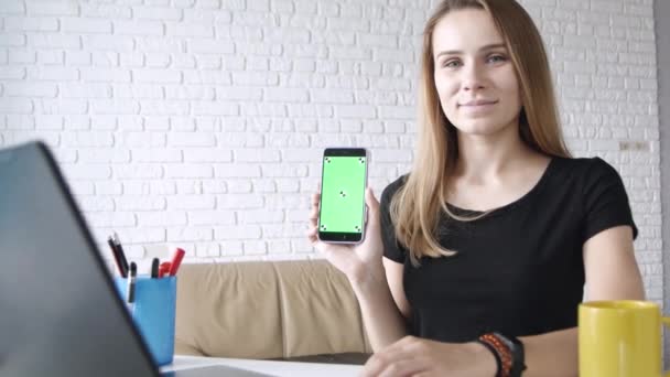 年轻的白种人长发女孩的肖像微笑着显示绿色屏幕的智能手机相机在家庭办公室右手 — 图库视频影像