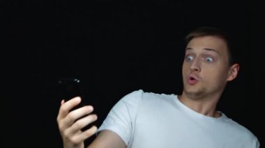 Genç beyaz adam şaşırttı, vay söyleyerek, telefon ekranına bakarak, piyango kazanmak. Portreyi kapat. Çok mutlu ve sevinen satış erkek siyah arka plan üzerinde duruyor. Başarı kavramı