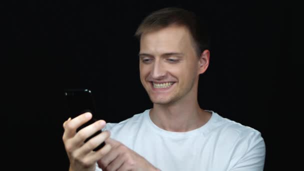 Leende Ung Kaukasiska Man Vit Shirt Med Smartphone Över Svart — Stockvideo
