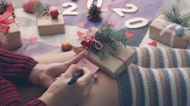 居心地の良い家の屋内で彼女の膝にピンクのラベルにクリスマスカードを書く若い女性 若い女の子が新年のはがきに署名します お父さんに手書きの碑文へ — ストック動画