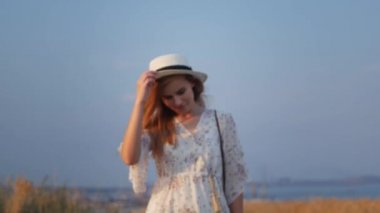 Beyaz elbise alanında yürüyüş portre genç güzel gülümseyen kadın yakın yukarı, onu hasır şapka alır, saçlarını düzeltir ve kameraya bakıyor. Doğa yavaş hareket video.