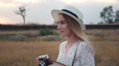 Beyaz elbise genç güzel kız arka plan günbatımı üzerinde, Vintage kamera ile alanda duruyor. Çayır üzerinde retro kamera ile fotoğraf çekmek hasır şapka neşeli kadın. Yavaş hareket, yakın çekim. 