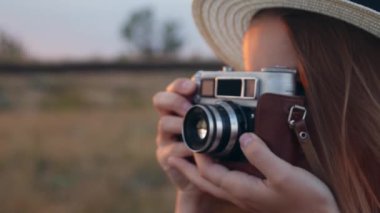 Beyaz elbise genç güzel kız arka plan günbatımı üzerinde, Vintage kamera ile alanda duruyor. Çayır üzerinde retro kamera ile fotoğraf çekmek hasır şapka neşeli kadın. Yavaş hareket, yakın çekim. 