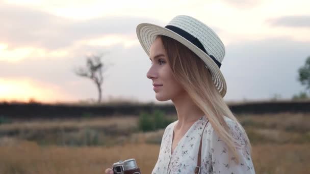白いドレスを着た若い美しい少女は 背景の夕日に ヴィンテージカメラでフィールドに立っています 牧草地でレトロなカメラで写真を撮るウィッカーハットの陽気な女性 スローモーション クローズアップ — ストック動画