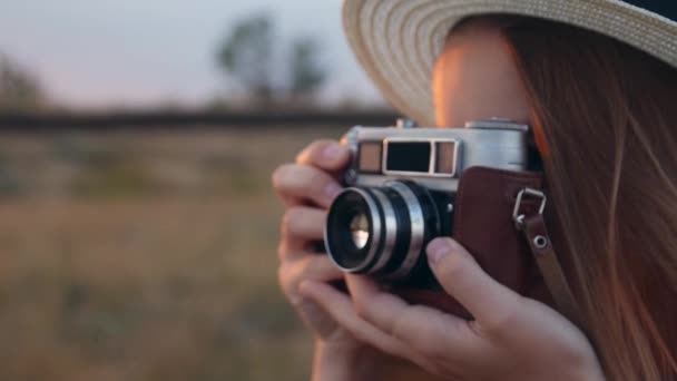 年轻的美丽的女孩在白色礼服站在田野与老式相机 背景日落 穿着柳条帽子的欢快的女人在草地上用复古相机拍照 慢动作 — 图库视频影像