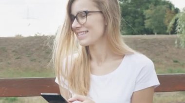 Mutlu kız öğrenci bir bankta oturan bir şehir parkında bir akıllı telefon kullanarak. Gözlüklü genç beyaz kadın, güler yüzlü kullanım uygulaması, uzun sarı saçlı, yakından. 