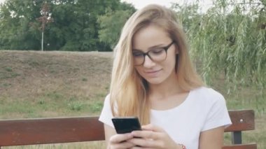 Mutlu kız öğrenci bir bankta oturan bir şehir parkında bir akıllı telefon kullanarak. Gözlüklü genç beyaz kadın, güler yüzlü kullanım uygulaması, uzun sarı saçlı, yakından. 