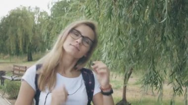 Gözlüklü rahat genç kadın beyaz bir tişört giyerek müzik dinliyor ve şehir parkında yürüyor. Gerçek zamanlı çekim kurma izleme. Açık havada yürüyen gülümseyen bayan müzik dinliyor. 