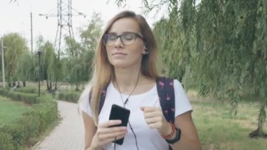 Gözlüklü rahat genç kadın beyaz bir tişört giyerek müzik dinliyor ve şehir parkında yürüyor. Gerçek zamanlı çekim kurma izleme. Açık havada yürüyen gülümseyen bayan müzik dinliyor. 