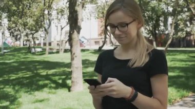 Akıllı telefonda uygulamayı kullanan, cep telefonunda gülümseyen ve mesaj veren genç güzel kadının portresi. Şehir parkında gözlüklü beyaz kadın yürüyor.