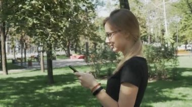 Akıllı telefonda uygulamayı kullanan, cep telefonunda gülümseyen ve mesaj veren genç güzel kadının portresi. Şehir parkında gözlüklü beyaz kadın yürüyor.
