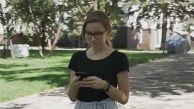 Gözlüklü, kameraya doğru yürüyen, telefonu kontrol eden ve parkta iyi haberleri kutlayan genç beyaz bir kadın.