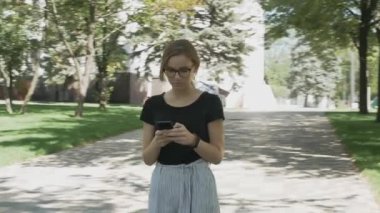 Gözlüklü, kameraya doğru yürüyen, telefonu kontrol eden ve parkta iyi haberleri kutlayan genç beyaz bir kadın.