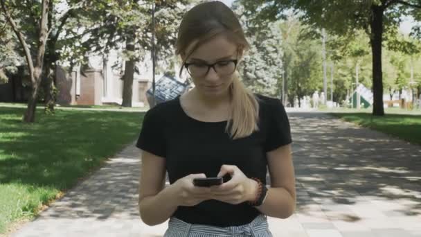 スマートフォンでアプリを使用して若い美しい女性の肖像画 笑顔と携帯電話でテキストメッセージ 市立公園を歩く眼鏡をかけた白人女性 — ストック動画