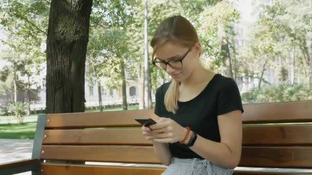 黒い携帯電話のメガネでかなり白人の女性が使用し 市内の公園のベンチに座ります — ストック動画