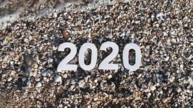 2020 yılı kumdaki sayılar, deniz kıyısındaki taş ve kabukların arasında bir deniz dalgası tarafından yıkanır. Yeni Yıl 2020 kavramı geliyor - bir kum plajda rakamlar. Yeni yıl tatili. Close. Üstte görüntü. 