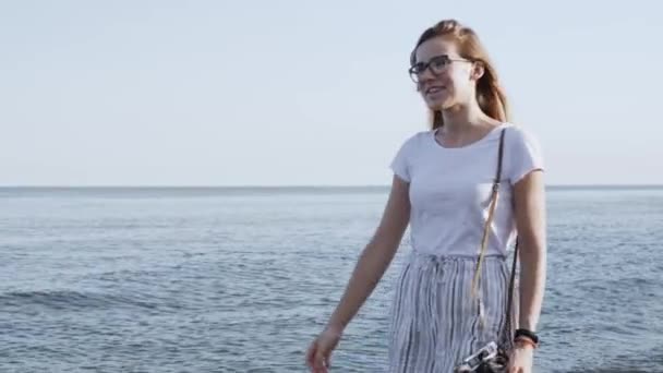 夕日を背景に古いレトロなカメラで海浜を歩く白いショートパンツの若い美しい女性 クローズアップ ビーチで自然と日光を楽しむ気楽な女性 — ストック動画