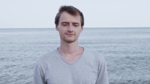 灰色のTシャツを着て夕暮しの穏やかな海辺のビーチで自信を持って微笑む魅力的な魅力的な若い白人男性の肖像画 — ストック動画