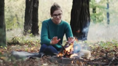 Soğuk günbatımında kamp ateşinin yanında oturan ve sonbaharda ormanda ellerini ısıtan beyaz bir kadın.. 