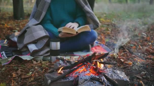 秋の寒い日没にキャンプファイヤーの近くで本を読んでいるかなり笑顔の女性 健康的なアクティブライフスタイルを生きる人々 閉じろ — ストック動画