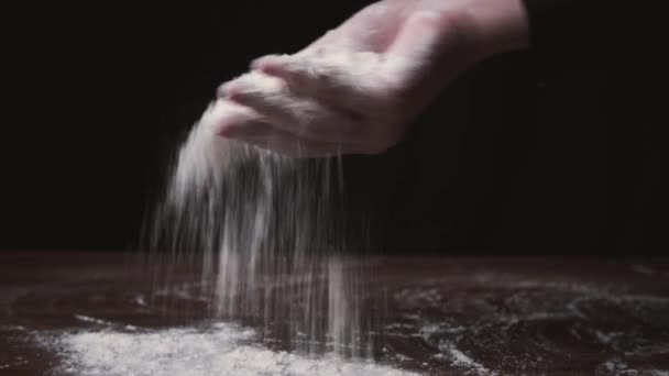 Harina cayendo de la mano. Primer plano de la fabricación y prepara la harina para la panadería casera. La mano de la mujer arrojando harina. Cocinar espolvorea harina sobre mesa de madera sobre fondo negro . — Vídeos de Stock
