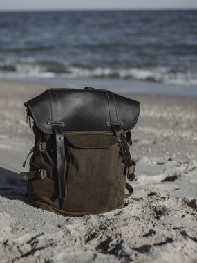 Hipster hiker tourister khaki backpack close up on background blue sea ocean horizon on sand beach, blurred panoramic seascape blank, traveler relax holiday concept, sunlight view in trip vacation clipart