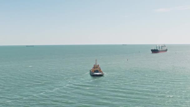 Εναέρια drone βίντεο του κίτρινου ρυμουλκού πηγαίνει σε ένα μεγάλο φορτηγό πλοίο για να μεταφερθεί στην αποβάθρα. Το φορτηγό πλοίο φορτηγίδα και ρυμουλκό πλέουν για να συναντηθούν στο λιμάνι του λιμανιού. Αεροφωτογραφία. — Αρχείο Βίντεο