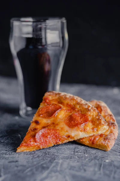 Slices of pepperoni pizza on dark black board, traditional italian pizza, glass of cola.