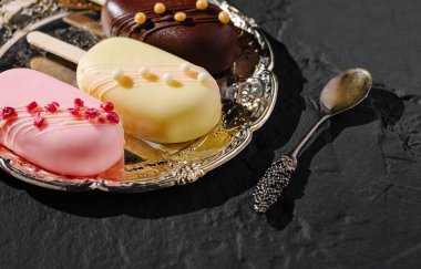 Pembe, sarı ve koyu çikolata aromalı renkli gurme buzlar şık bir şekilde gümüş tepside sunuluyor. Yakınlarda bir kaşık oturuyor, bu lezzetli tatları tatmaya hazır..