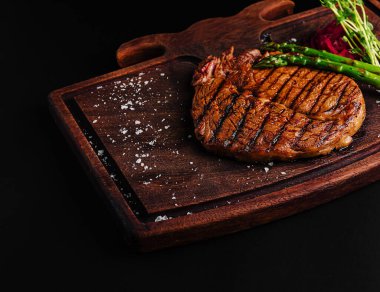 Güzel bir ızgara biftek ahşap bir kesme tahtasına konur. Yanında kuşkonmaz ve sos serpiştirilir. Dekor, zengin, kavrulmuş dokuyu ve davetkar aromayı ön plana çıkarıyor..