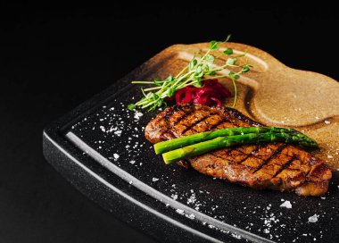 Güzel bir ızgara biftek parlak siyah bir tabakta sunulur. Taze kuşkonmaz ve mikro yeşiller yemek sanatını ön plana çıkaran renkli bir garnitürle birlikte tabağı süslüyor..