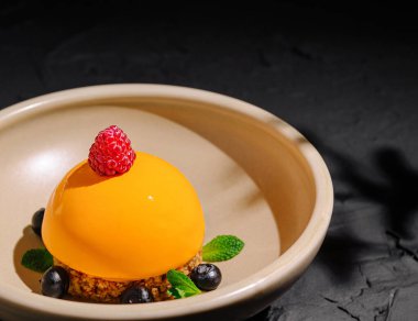 Canlı turuncu bir tatlı bej bir tabağın üzerinde, ahududu ve yaban mersiniyle süslenmiş olarak oturur. Yaradılış, taze nane ile çevrili pürüzsüz bir doku sergiliyor, zarif bir sunum sunuyor..