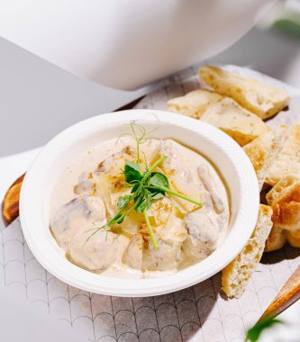 Lezzetli kremalı bir tavuk tabağı, yanında taze pişmiş ekmek dilimleriyle beyaz bir kasede oturur. Dekorasyonda zarif dekorlu temiz ve modern bir yemek ortamı yer alıyor..