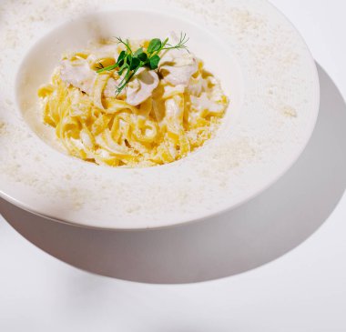 Çok güzel bir kaplanmış kremalı makarna dairesel bir tabakta oturur. Etrafı hafif ışıklandırma ve yakınlardaki yeşilliklerin nazik gölgeleriyle çevrilidir. Bu da yemek pişirme deneyimi yaratır..