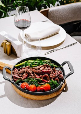 Yanında canlı kuşkonmaz ve kiraz domatesleri olan ızgara biftek tabağı şık bir yemek masasında şarapla zarif bir şekilde sunulur..