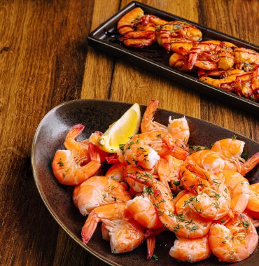 Koyu bir tabakta limon dilimi ile lezzetli bir karides çeşidi sunulur. Siyah tepside ızgara karides, ahşap bir masada yaratılan yemek atmosferine tat katar..