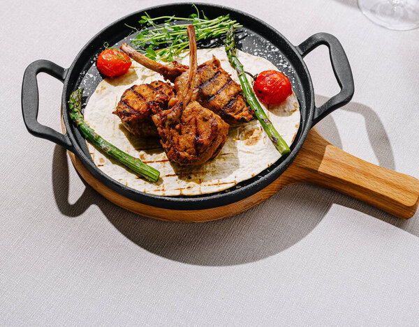 Juicy grilled lamb chops are beautifully presented on a skillet. Accompanied by asparagus and cherry tomatoes, this dish is set on a sophisticated dining table with elegant glassware.