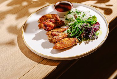 Lezzetli ızgara tavuk kanatları, canlı bir salatanın yanında beyaz bir tabakta, yapraklı yeşillikler ve keskin bir sosla birlikte, hepsi de ahşap bir masaya yerleştirilmiş..