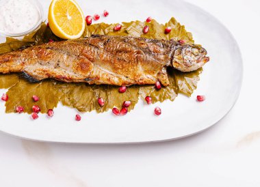 Izgara balık, üzerinde üzüm yaprakları bulunan, nar tohumları ve limon dilimleriyle süslenmiş, canlı ve iştah açıcı bir tabakta sunulur..