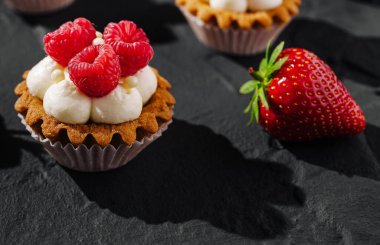 Üzerinde krema ve taze ahududu bulunan tart tabanlı güzel bir tatlı canlı bir çileğin yanında duruyor. Ayarlar koyu dokulu bir yüzey gösteriyor..