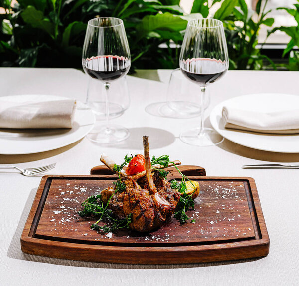A beautifully plated lamb dish garnished with herbs is presented on a wooden board. Two elegant wine glasses filled with red wine accompany the meal, creating a sophisticated dining atmosphere.