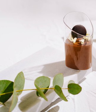 Yumuşak bir çikolatalı tatlı zarif bir şekilde çikolata küresi ve çiçek aksanıyla kaplanmış modern bir bardakta sunulur. Beyaz bir masa örtüsüne konur ve yanında yeşil bir yaprak vardır..