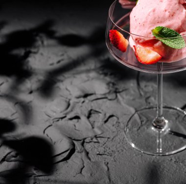 Lezzetli bir çilek şerbeti zarif bir bardakta sunulur, taze nane yaprakları ve dilimlenmiş çileklerle süslenir, dokulu siyah bir yüzey üzerinde dinlenerek canlı rengini arttırır..