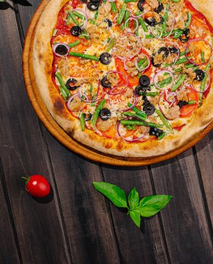 Zeytin, soğan ve yeşil biberli taze pişmiş sebze pizzası ahşap bir masada oturur. Olgun bir domates ve taze fesleğen, ayara canlı renkler ekleyerek eşlik eder..