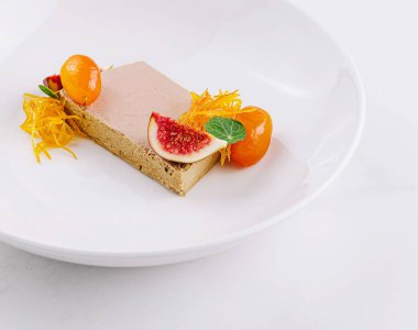 Beyaz bir tabakta incir, kumkat ve yenilebilir çiçeklerle süslenmiş kaz ciğeri dilimleri servis edilir.