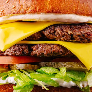 Duble çizburger iki köfte, erimiş peynir ve taze sebzelerle üst üste dizilmiştir. Canlı renkler marul, domates ve turşunun tazeliğini vurguluyor..