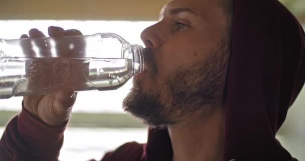 Hombre adulto joven con sudadera con capucha agua potable descansando durante el entrenamiento deportivo de fitness. Grunge entrenamiento urbano industrial. 4k video en cámara lenta — Vídeos de Stock